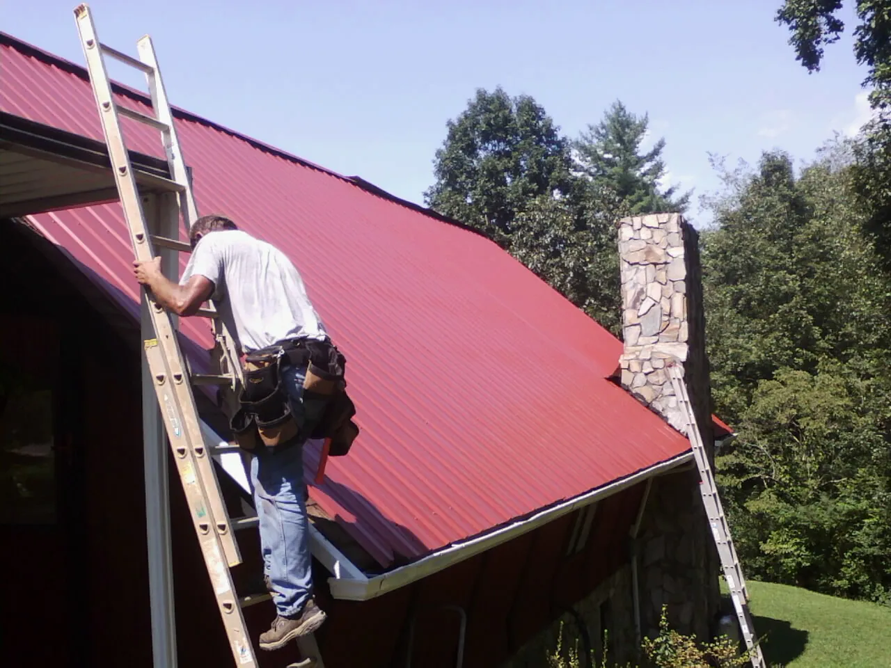 Roofer on a ladder beside a red metal roof and stone chimney — Soffit & Fascia Repair by Roofing Brothers in Wolcottville